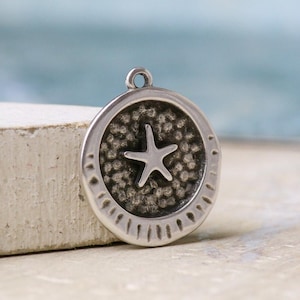 Puede incluir: Un colgante circular de metal plateado con un diseño de estrella de mar en relieve en el centro. El fondo del colgante tiene un acabado texturizado, oscuro y granulado. El borde exterior es liso con líneas radiales grabadas en la parte inferior.