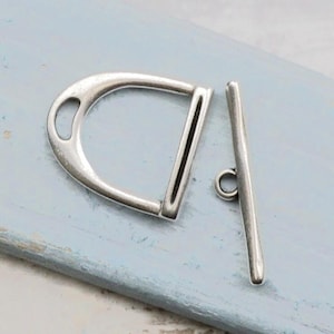 Op de afbeelding: Een zilverkleurige metalen toggle-sluiting bestaande uit twee afzonderlijke delen: een D-vormige lus met een kleine ronde opening aan de bovenkant en een rechte staaf met een centrale ring, ontworpen voor het sluiten van sieraden of kleding.