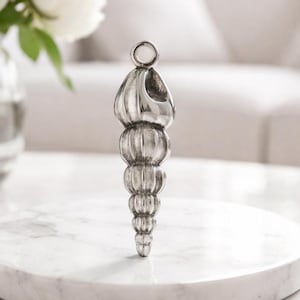 Op de afbeelding: Een zilverkleurige flesstopper in de vorm van een zeeschelp. De stopper heeft een ring aan de bovenkant en een reeks geribbelde, bolvormige secties die taps toelopen naar een punt. Het wordt weergegeven op een wit marmeren oppervlak.