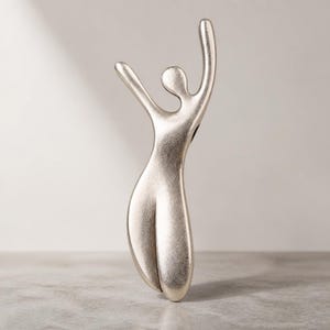 Peut inclure: Une sculpture abstraite argentée représentant une silhouette humaine avec une finition métallique lisse. La figure est représentée dans une pose dynamique et verticale, un bras levé et l'autre étendu, avec une forme corporelle incurvée et allongée.