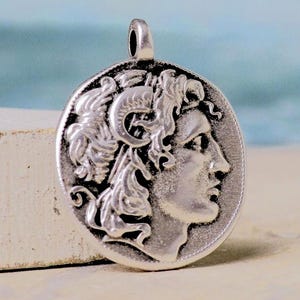 Op de afbeelding: Een cirkelvormige zilverkleurige hanger met een reli&euml;f van een profiel van een figuur met krullend haar en een hoornachtig detail op het hoofd. Het metalen oppervlak heeft een donkere geoxideerde afwerking om het ontwerp te benadrukken.