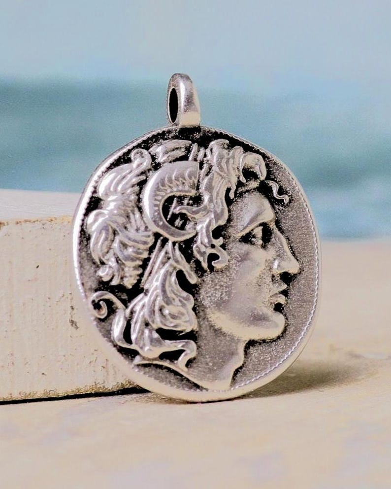 Op de afbeelding: Een cirkelvormige zilverkleurige hanger met een reli&euml;f van een profiel van een figuur met krullend haar en een hoornachtig detail op het hoofd. Het metalen oppervlak heeft een donkere geoxideerde afwerking om het ontwerp te benadrukken.