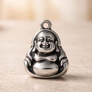 Puede incluir: Un colgante de metal plateado pulido con forma de figura de Buda sentado y sonriente. El dije presenta un diseño redondeado y suave con un pequeño lazo circular en la parte superior para colgar, mostrando un acabado reflectante y detalles.