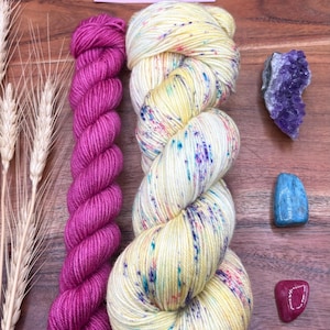 May include: An overhead shot of yarn skeins, crystals, and wheat stalks on a wooden surface. The yarn is in shades of yellow, pink, and purple. The crystals are amethyst, blue, and red. A pink label reads "Potions & Purls - Hand Dyed Yarn."
