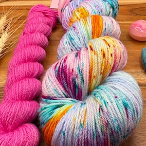 May include: A collection of colorful yarn skeins. One skein is bright pink, while the others are a mix of blue, pink, yellow, and orange. The yarn is arranged on a wooden surface, suggesting a craft or hobby theme.
