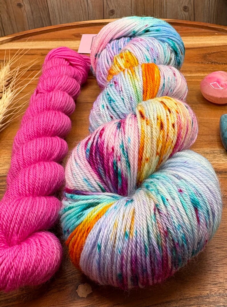 May include: A collection of colorful yarn skeins. One skein is bright pink, while the others are a mix of blue, pink, yellow, and orange. The yarn is arranged on a wooden surface, suggesting a craft or hobby theme.