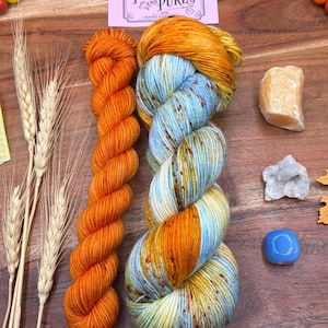 May include: Two skeins of yarn, one solid vibrant orange and one variegated with light blue, yellow, and orange speckles. A pink rectangular label with the text Potions and Purls is positioned above the yarn, accompanied by dried wheat stalks.