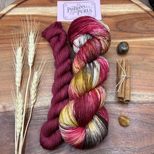 May include: A skein of hand-dyed yarn in shades of burgundy, gold, and cream, next to a smaller skein of solid burgundy yarn. The image also includes wheat stalks, cinnamon sticks, and a small card with the text "Potions & Purls".