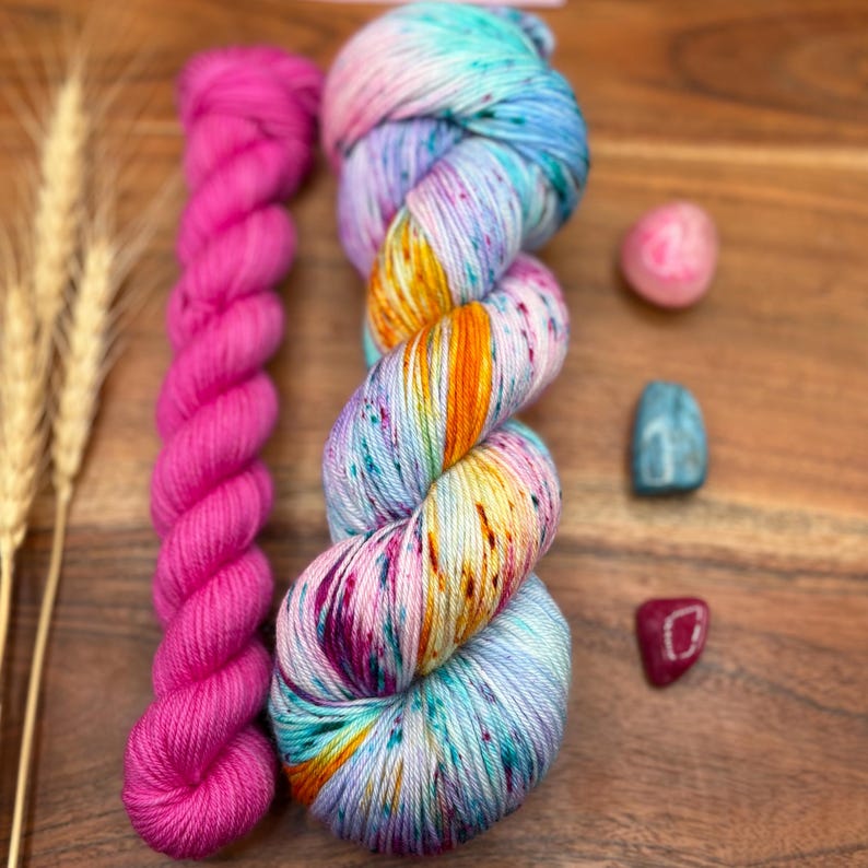 May include: A skein of colorful yarn with shades of blue, orange, and pink, next to a bright pink yarn and three small gemstones. The yarn is on a wooden surface, with wheat stalks in the background.