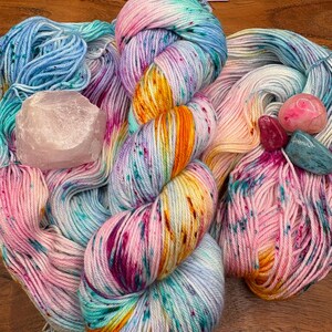 May include: Colorful hand-dyed yarn skeins in shades of blue, pink, yellow, and purple. A large, light pink crystal and several small polished gemstones in pink and blue are also present. The yarn is arranged in a visually appealing display.