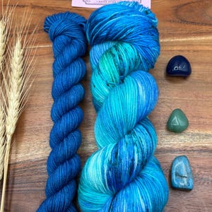 May include: Two skeins of hand-dyed yarn in shades of blue and teal, with a small skein of solid blue yarn. The image also includes three small gemstones and a card with the text "Potions & Purls - Hand Dyed".
