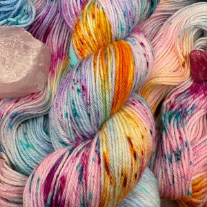 May include: A close-up of colorful hand-dyed yarn in shades of pink, blue, yellow, and purple. The yarn is speckled with darker hues and has a soft, textured appearance. A pink crystal is visible in the background. This yarn is perfect for knitting and crochet projects.