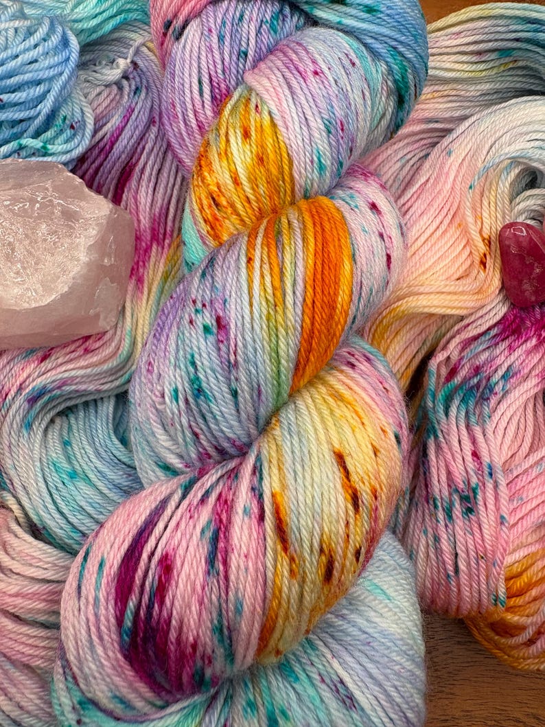 May include: A close-up of colorful hand-dyed yarn in shades of pink, blue, yellow, and purple. The yarn is speckled with darker hues and has a soft, textured appearance. A pink crystal is visible in the background. This yarn is perfect for knitting and crochet projects.