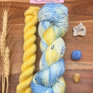 May include: Two skeins of hand-dyed yarn in yellow and blue hues, with a small crystal and two polished stones. The yarn is labeled "Potions and Purls - Hand Dyed Yarn". The background is a wooden surface, with a few stalks of wheat.