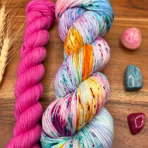 May include: A skein of vibrant, multicolored yarn with shades of blue, pink, orange, and purple, next to a bright pink skein. The image also includes small gemstones and a card with the text "Potions & Purls".