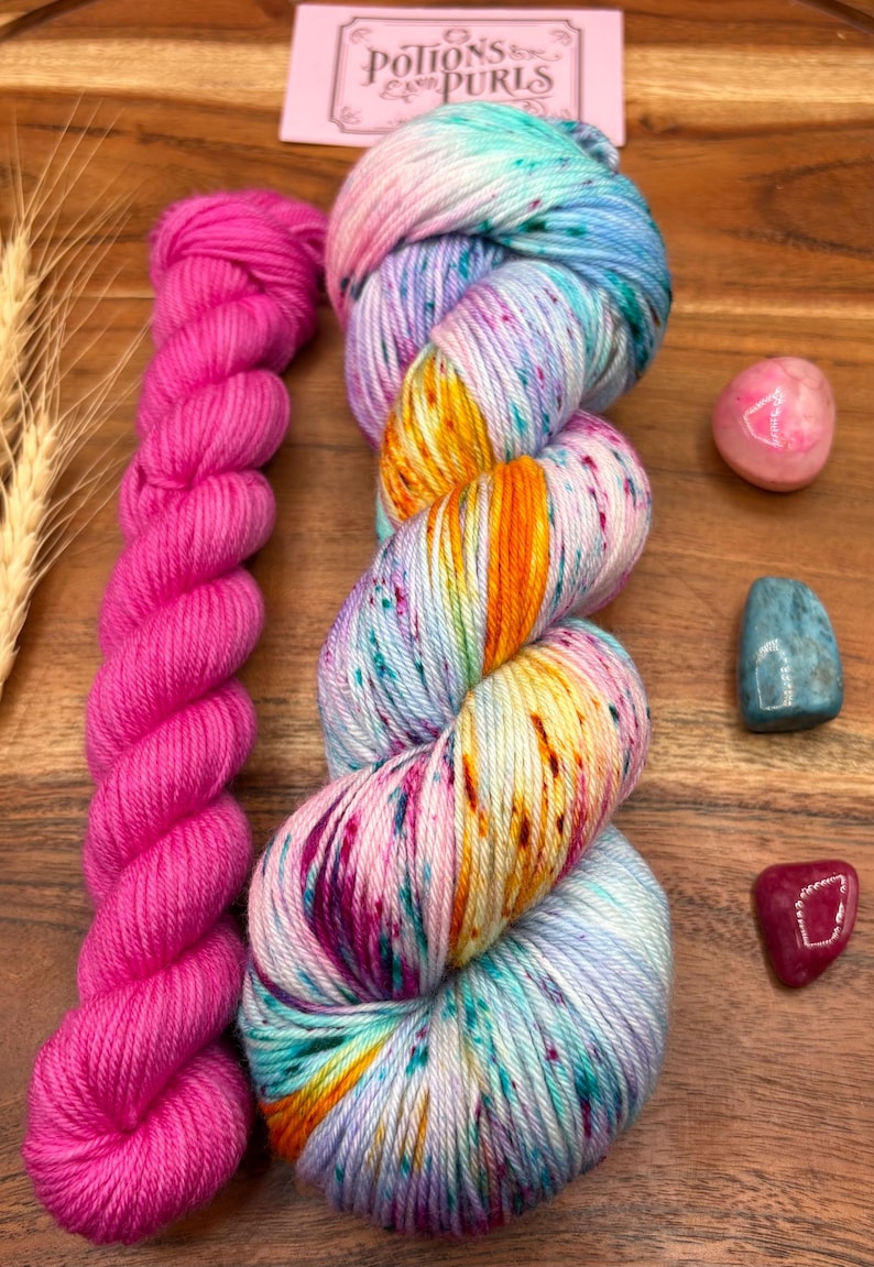 May include: A skein of vibrant, multicolored yarn with shades of blue, pink, orange, and purple, next to a bright pink skein. The image also includes small gemstones and a card with the text "Potions & Purls".