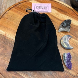 May include: A black velvet drawstring bag with a pink tag that reads "Potions and Purls". The bag is next to a smoky quartz crystal, a silver crescent moon, and a purple amethyst cluster. The items are on a wooden surface.