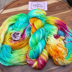 May include: A skein of hand-dyed yarn in vibrant colors of yellow, green, turquoise, orange, and pink. A small geode and several polished stones are placed near the yarn. A small sign in the background reads "Potions and Purls".