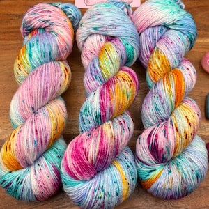 May include: Three skeins of colorful yarn in a variety of colors, including pink, yellow, blue, and green. The yarn is speckled with darker flecks, creating a textured appearance. The yarn is arranged in a twisted pattern.