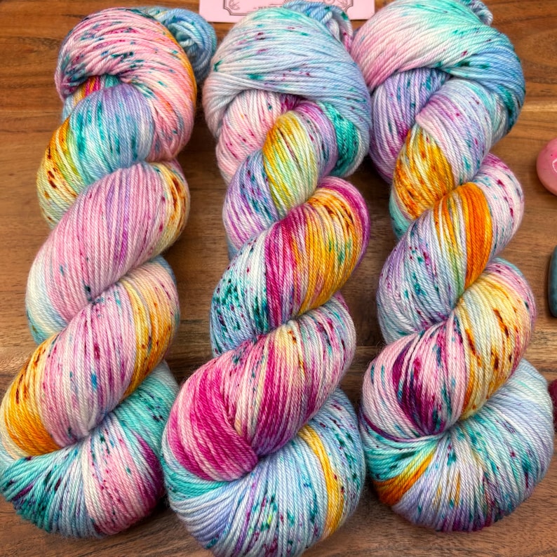 May include: Three skeins of colorful yarn in a variety of colors, including pink, yellow, blue, and green. The yarn is speckled with darker flecks, creating a textured appearance. The yarn is arranged in a twisted pattern.