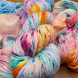 May include: A collection of colorful yarn skeins in shades of pink, blue, orange, and purple. The yarn is speckled with various colors and is arranged on a wooden surface. Also included are crystals.