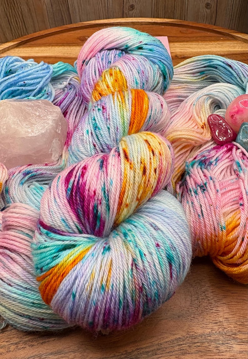 May include: A collection of colorful yarn skeins in shades of pink, blue, orange, and purple. The yarn is speckled with various colors and is arranged on a wooden surface. Also included are crystals.
