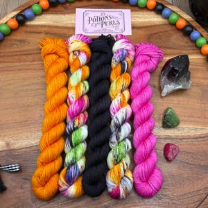 May include: Five skeins of yarn arranged vertically, featuring solid orange, variegated orange-pink-green, solid black, variegated orange-purple-white, and solid hot pink. A rectangular tag reads Potions and Purls. Three crystals are placed to the right.