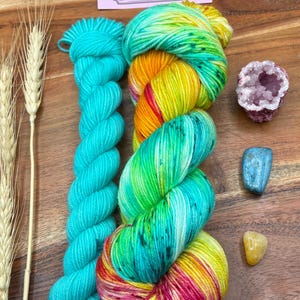 May include: A still-life composition featuring hand-dyed yarn in vibrant colors, including turquoise, yellow, and pink. The image also includes a small geode, crystals, and wheat stalks, with a label that reads "Potions & Purls - Hand Dyed Yarn."