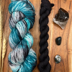 May include: Two skeins of yarn, one teal and black variegated and one black. There are also four small stones, two green, one clear and one black.