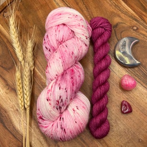 May include: Two skeins of yarn, one pink with white speckles and one deep burgundy. The yarn is arranged on a wooden surface with two pink heart-shaped stones and a silver crescent moon charm.
