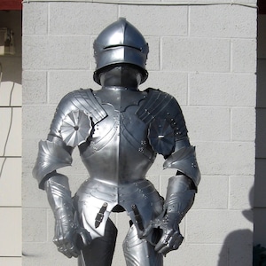 May include: A full suit of silver-colored medieval armor, including a helmet, chest plate, arm and leg protection, and gauntlets. The armor is displayed on a dark wooden stand against a white brick wall.