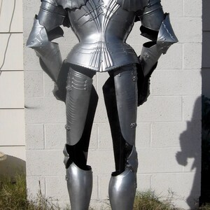 May include: A full suit of silver-colored medieval armor, including a helmet, chest plate, arm and leg protection. The armor is displayed on a dark wooden stand against a white brick wall. The armor is highly detailed with intricate designs.