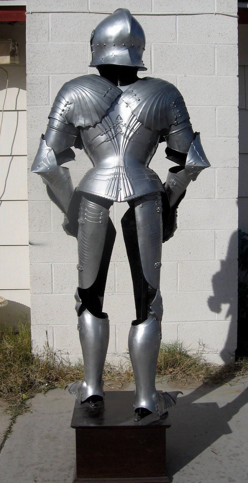 May include: A full suit of silver-colored medieval armor, including a helmet, chest plate, arm and leg protection. The armor is displayed on a dark wooden stand against a white brick wall. The armor is highly detailed with intricate designs.