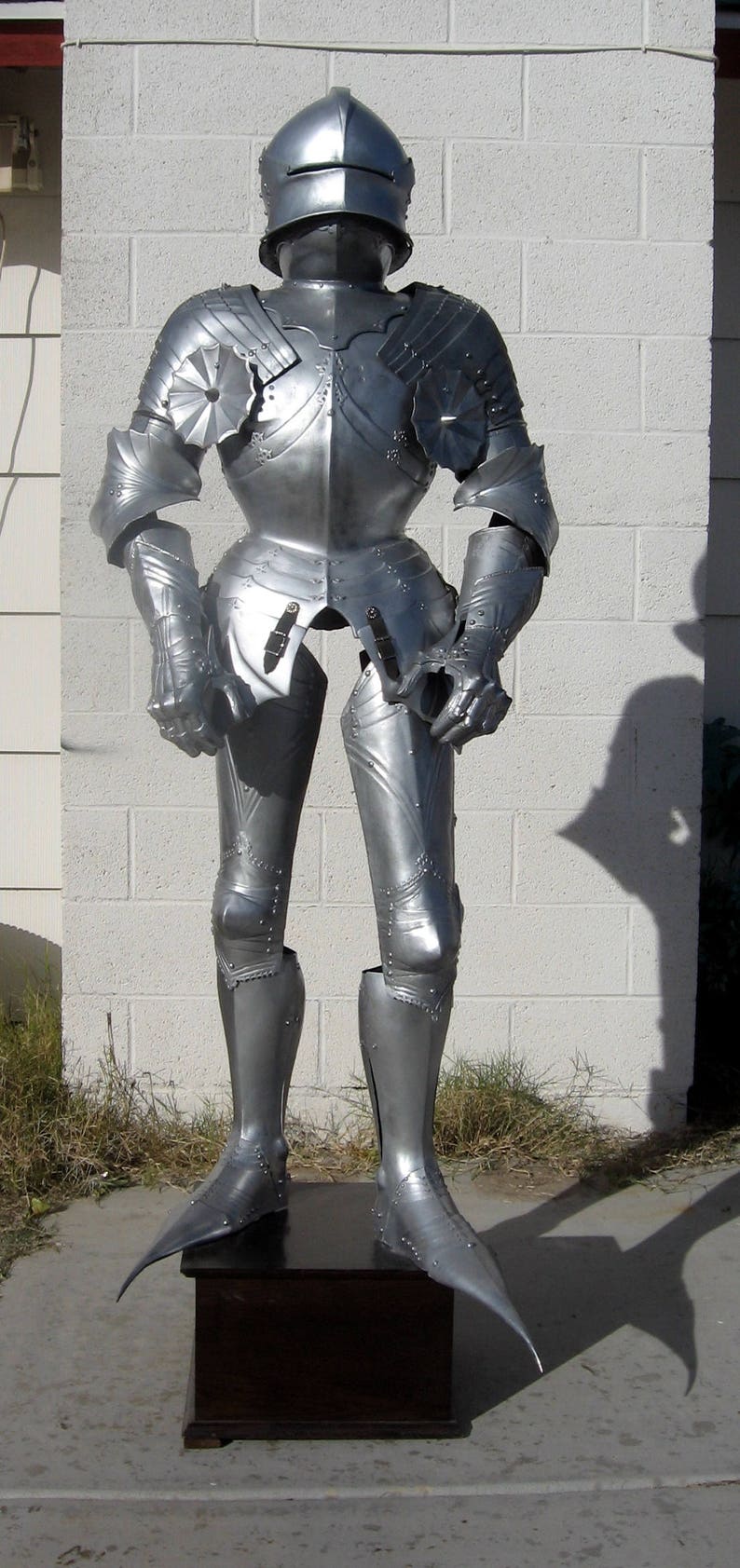 May include: A full suit of silver-colored medieval armor, including a helmet, chest plate, arm and leg protection, and gauntlets. The armor is displayed on a dark wooden stand against a white brick wall.
