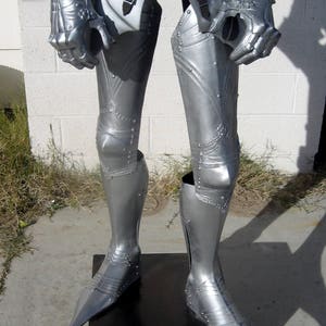 May include: Silver-colored medieval armor, including articulated gauntlets, greaves, and cuisses. The armor is riveted and detailed, with pointed sabatons. The legs are positioned on a dark wooden stand, suggesting a display or costume.