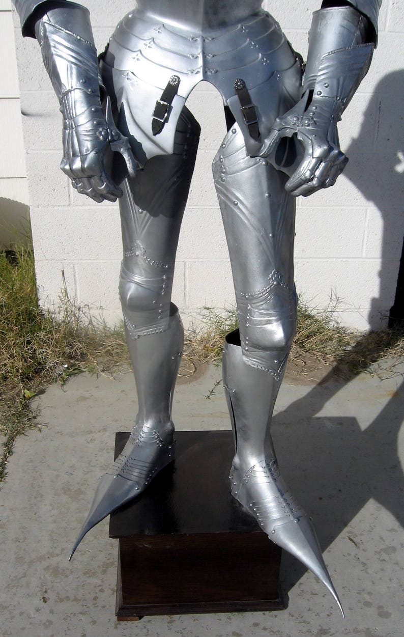 May include: Silver-colored medieval armor, including articulated gauntlets, greaves, and cuisses. The armor is riveted and detailed, with pointed sabatons. The legs are positioned on a dark wooden stand, suggesting a display or costume.