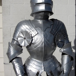 May include: A full suit of silver-colored medieval armor, including a helmet, chest plate, and arm and leg protection. The armor is highly detailed with decorative elements and buckles. The suit is standing upright against a white brick wall.