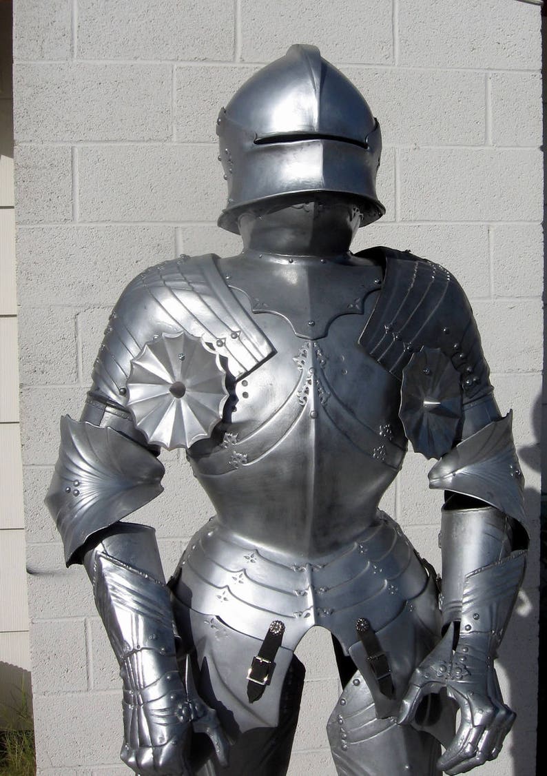 May include: A full suit of silver-colored medieval armor, including a helmet, chest plate, and arm and leg protection. The armor is highly detailed with decorative elements and buckles. The suit is standing upright against a white brick wall.