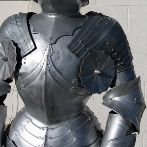 May include: A detailed shot of a silver-colored suit of armor. The armor features articulated plates, decorative rivets, and ornate embellishments. The design includes shoulder pauldrons, chest plates, and arm protection, showcasing historical craftsmanship.