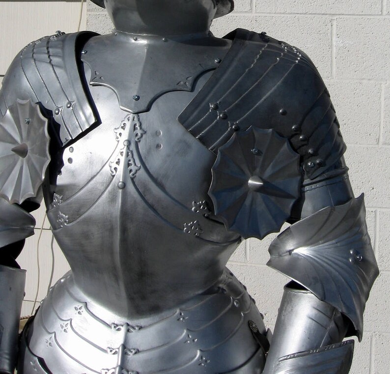 May include: A detailed shot of a silver-colored suit of armor. The armor features articulated plates, decorative rivets, and ornate embellishments. The design includes shoulder pauldrons, chest plates, and arm protection, showcasing historical craftsmanship.