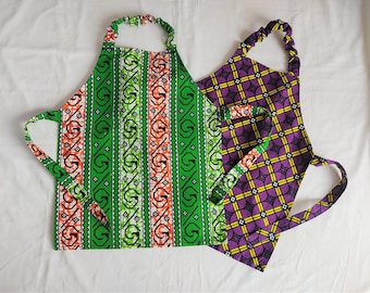 Könnte beinhalten: Zwei Schürzen mit farbenfrohen, afrikanisch inspirierten Mustern. Eine Schürze hat ein grünes, orangefarbenes und weißes Design, die andere ein geometrisches Muster in Lila und Gelb. Beide haben verstellbare Nacken- und Taillenbänder.