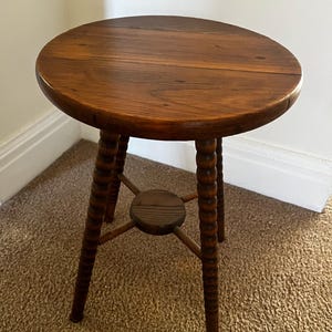 May include: A small, round wooden stool with a dark brown finish. The stool has a circular seat and three turned legs connected by a lower support. The legs have a decorative, ribbed design.