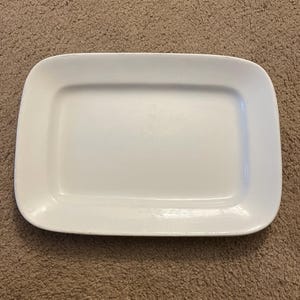 May include: A rectangular white ceramic serving platter with rounded corners and a smooth, glossy finish. The dish features a slightly recessed center area and a wide, flat rim that extends to the edges, creating a clean and simple design.