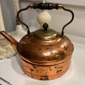 May include: A vintage copper tea kettle with a wooden handle and brass accents. The kettle has a worn, aged patina.