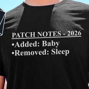 Puede incluir: Camiseta negra con el texto "PATCH NOTES - 2026" y la lista con viñetas "Added: Baby" y "Removed: Sleep". Un logotipo que dice "-FROM- NERDS -WITH- LOVE" está en la esquina superior izquierda.