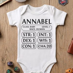 May include: White baby onesie with the name "ANNABEL" printed on the front, along with role-playing game stats. The onesie includes the text "CLASS: BABY, RACE: HUMAN, LEVEL: 1". Other items include dice, a dragon figurine, and a leather-bound journal.