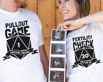 Puede incluir: Una pareja lleva camisetas blancas con un gráfico de dados y el texto "Pullout Game Critical Failure" y "Fertility Check Critical Hit". La mujer sostiene una imagen de ultrasonido.