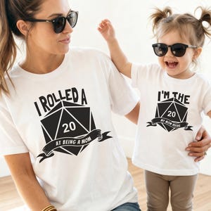 Puede incluir: Dos camisetas blancas con texto y gráficos negros. Una camiseta dice "I Rolled a 20 at Being a Mom" y la otra "I'm the 20 My Mom Rocks". Ambas camisetas presentan un gráfico de dado D20 negro.