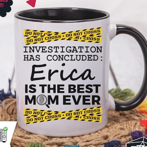 Puede incluir: Taza de cerámica blanca con asa y borde negros. La taza presenta el texto "INVESTIGATION HAS CONCLUDED: Erica IS THE BEST MOM EVER" con gráficos de cinta de precaución amarilla y negra. La taza es apta para lavavajillas y microondas.