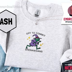 May include: Ash grey sweatshirt featuring the embroidered text "OFF TO COMMIT SHENANIGANS" and a cartoon frog in a wizard outfit. The sweatshirt has a crew neck and long sleeves. The brand name "GILDAN" is visible on the tag.